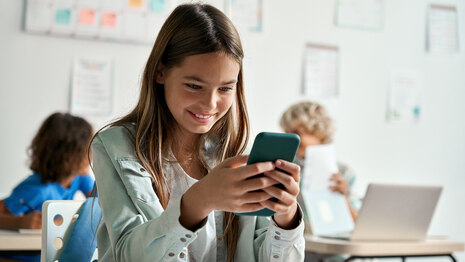 junges Mädchen in Klassenzimmer schaut lächelnd auf Smartphone