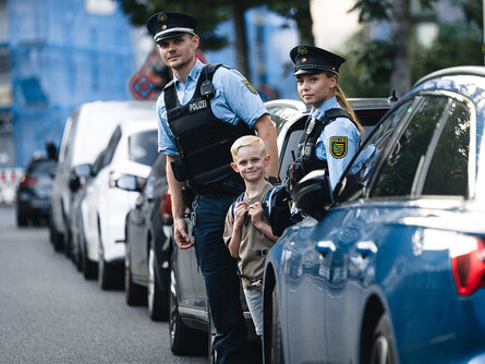 Die Schule hat begonnen: Ein Polizist, eine Polizistin und ein Schulkind stehen am Straßenrand zwischen geparkten Autos. Sie beobachten den Verkehr.