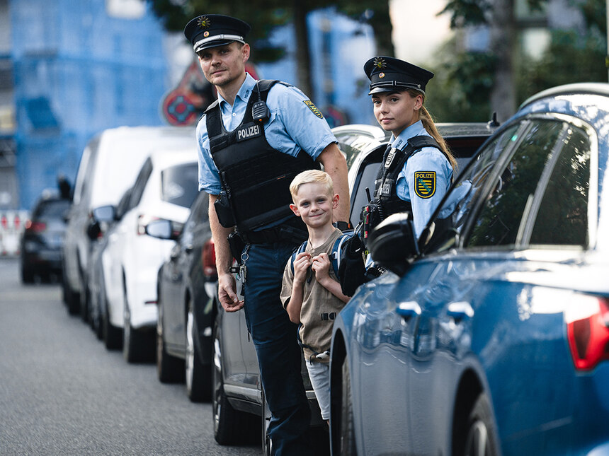 Die Schule hat begonnen: Ein Polizist, eine Polizistin und ein Schulkind stehen am Straßenrand zwischen geparkten Autos. Sie beobachten den Verkehr.