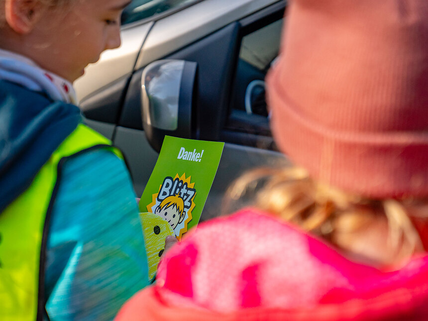 Blitz für Kids: Zwei Kinder werden von hinten gezeigt. Sie tragen eine gelbe Warnweste. Sie verteilen eine »grüne Karte« an einen Autofahrer. Die geöffnete Scheibe des Fahrzeugs ist zu sehen.