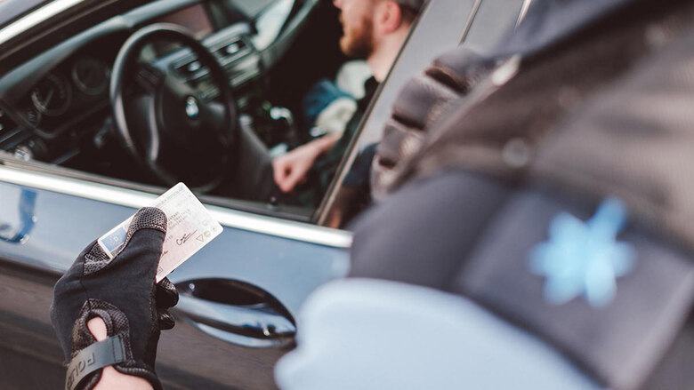 Verkehrskontrolle: Ein Polizist kontrolliert einen Autofahrer. Dieser ist hinter geöffneten Scheibe im Profil zu sehen. Der Polizist hält den Führerschein in der Hand.