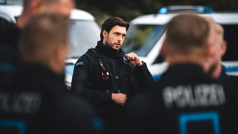 Ein Polizist der Bereitschaftspolizei in Einsatzuniform spricht in sein Funkgerät. Im Hintergrund sind Streifenwagen zu sehen, im Vordergrund das Team der Polizei Sachsen.