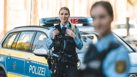 Zwei Polizistinnen stehen vor einem Streifenwagen. Dabei liegt der Fokus auf der Beamtin in blauer Polizeiuniform in der Mitte. Sie hat blonde, lange Haare und schaut in die Kamera.
