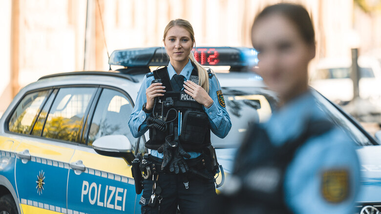 Zwei Polizistinnen stehen vor einem Streifenwagen. Dabei liegt der Fokus auf der Beamtin in blauer Polizeiuniform in der Mitte. Sie hat blonde, lange Haare und schaut in die Kamera.