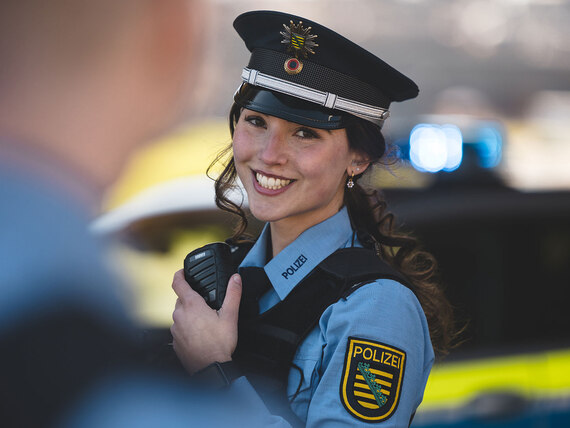 Eine junge Polizistin in der sächsischen Landeshauptstadt Dresden lächelt, während sie das Funkgerät bedient. Im Hintergrund ist unscharf ein Streifenwagen zu sehen.
