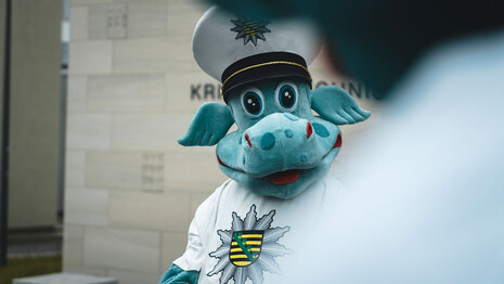 Kinderpolizist POLDI lächelt freundlich in die Kamera. Der grüne Polizeidino trägt eine Polizeimütze und hat für Kinder viele wichtige Tipps und zum Beispiel auch interessante Filme auf seiner Website.