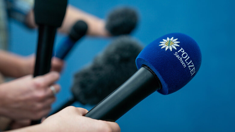 Mehrere Mikrofone sind vor blauem Hintergrund zu sehen. Auf einem ist das Logo und der Schriftzug »Polizei Sachsen« abgebildet.