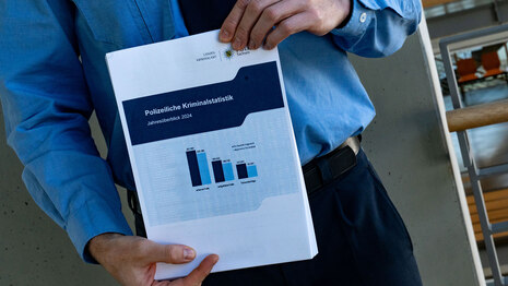 Ein Polizist in Uniform hält ein Dokument mit Graphen in der Hand. Das Dokument ist beschriftet mit: »Polizeiliche Kriminalstatistik – Jahresüberblick 2024«.