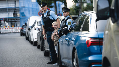 Eine Polizistin und ein Polizist in Uniform stehen zwischen parkenden Autos – mit einem Schulkind mit Schulrucksack. Die Beamtin und der Beamte sprechen mit dem Jungen, worauf beim sicheren Schulweg zu achten ist – nur eines der Themen in der Prävention.