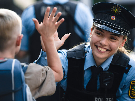 Bei der Polizei Sachsen gibt es einen vielfältigen Berufsalltag: Eine junge Polizistin und ein Kind mit Schulrucksack. Die Polizistin lächelt den Jungen an.