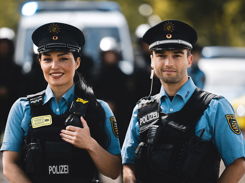 Polizei Sachsen wirbt um Nachwuchs: Eine Polizistin und und Polizist in blauer Polizeiuniform stehen auf einer Straße und lächeln in die Kamera. Hinter ihnen ist ein Polizeifahrzeug mit Blaulicht zu sehen.