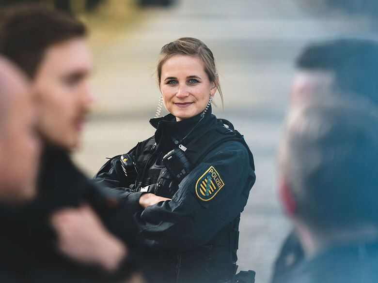 Eine junge Bereitschaftspolizistin steht in ihrer dunklen Uniform zwischen ihren Kollegen. Der Fokus ist auf sie gerichtet. Sie schaut in die Kamera.